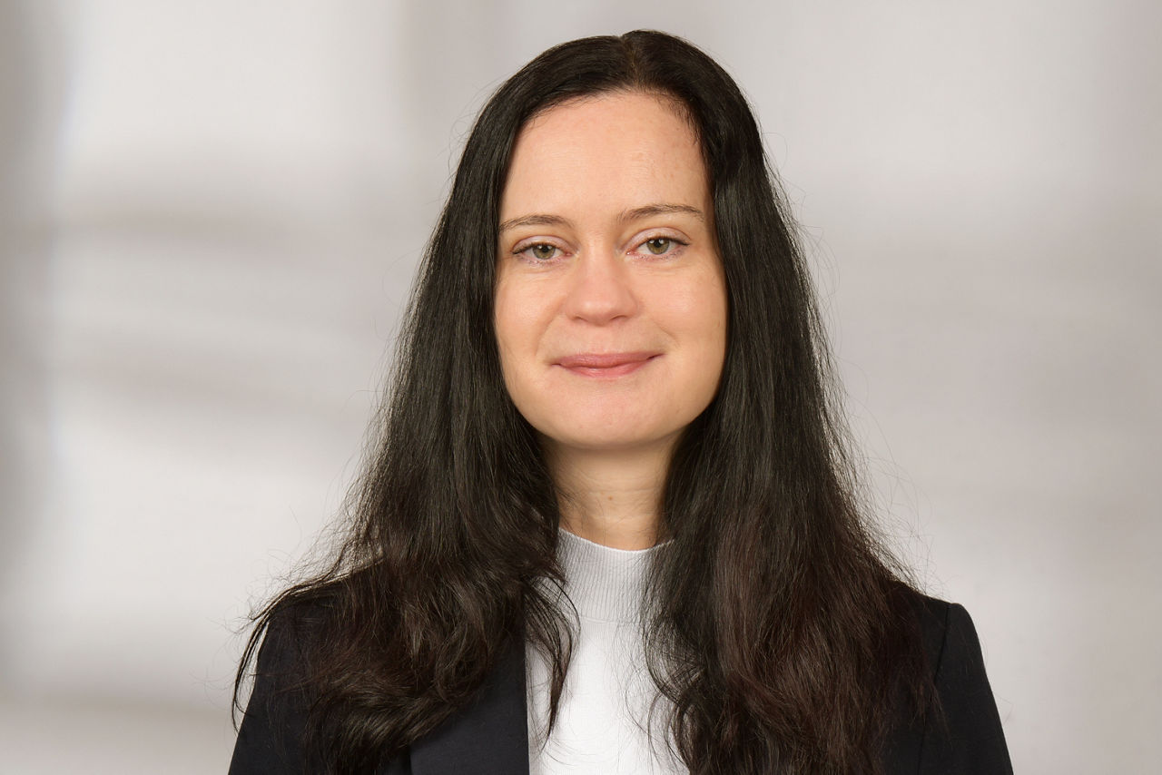 Person mit langen, dunklen Haaren trägt weißen Pullover und dunkle Jacke vor hellem Hintergrund