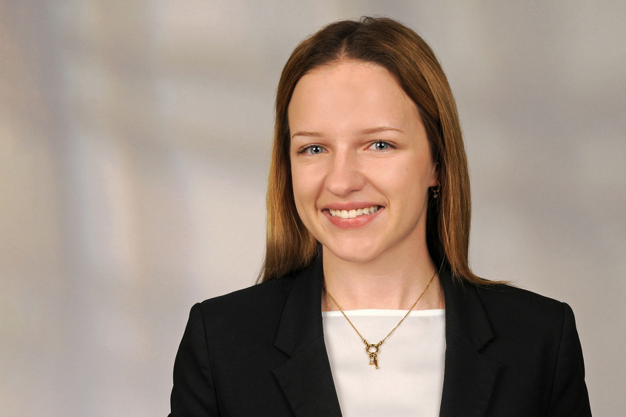 Frau mit schulterlangem braunem Haar trägt schwarzen Blazer, weiße Bluse und goldene Halskette mit Anhänger