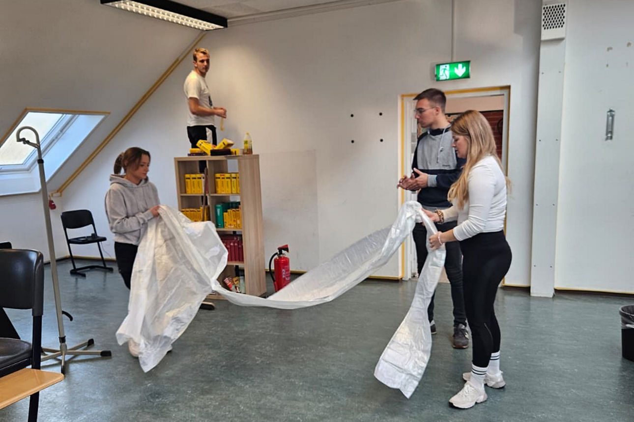 Zwei Frauen halten eine Abdeckplane zwischen sich. Ein Mann steht auf der Leiter und schaut zu den beiden Frauen. Hinter den beiden Frauen steht ein weiterer Mann.