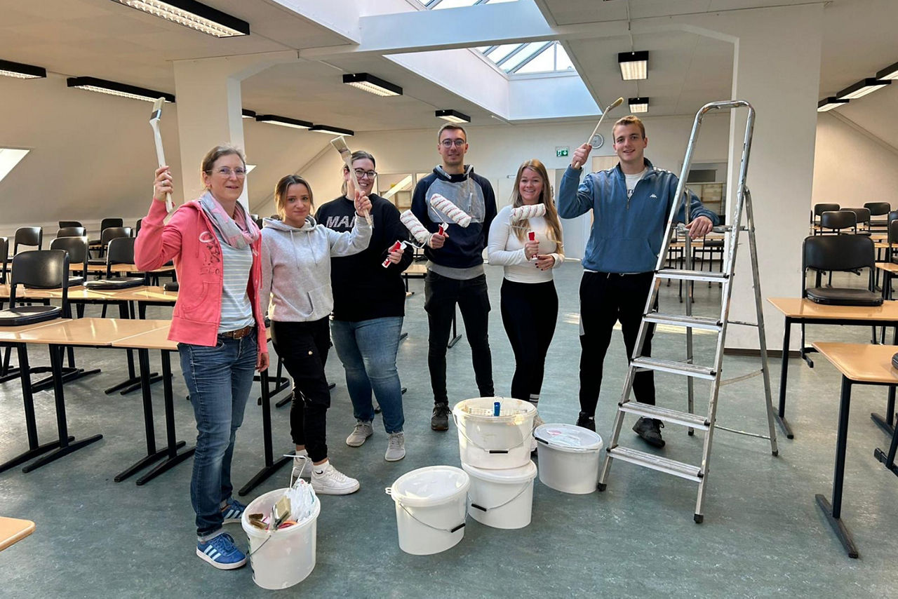 Unser Rent a Banker Team mit vier Frauen und zwei Männern halten Pinsel und Malrollen in der Hand. Vor ihnen stehen die Eimer mit der weißen Wandfarbe.