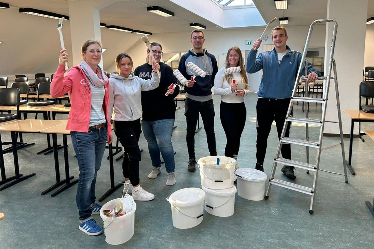 Unser Rent a Banker Team mit vier Frauen und zwei Männern halten Pinsel und Malrollen in der Hand. Vor ihnen stehen die Eimer mit der weißen Wandfarbe.