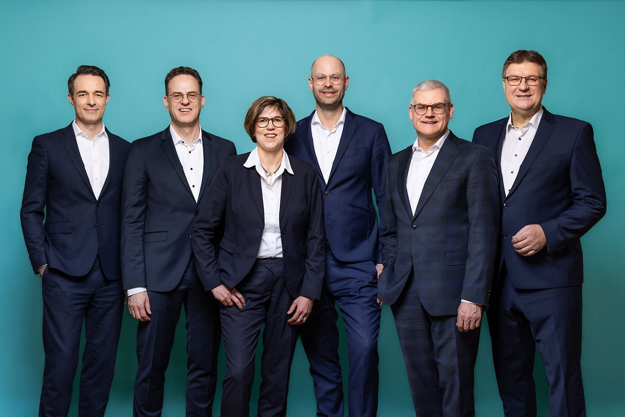 Der sechsköpfige Vorstand der neu entstandenen VR RegionalBank nebeneinanderstehend. Der Hintergrund ist petrolfarben.
