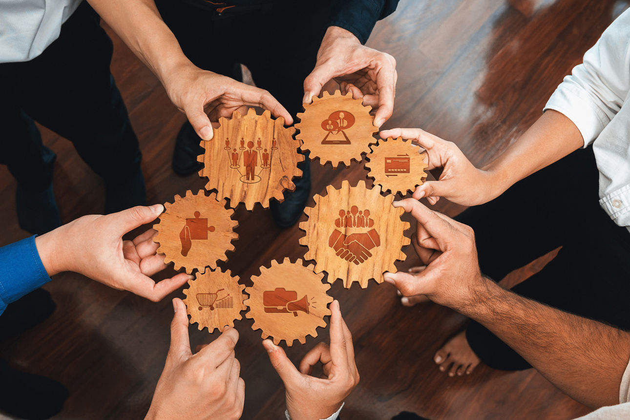 Hands holding wooden gears with business symbols represent teamwork and collaboration in a professional setting, highlighting unity and innovation within an organization. Amity