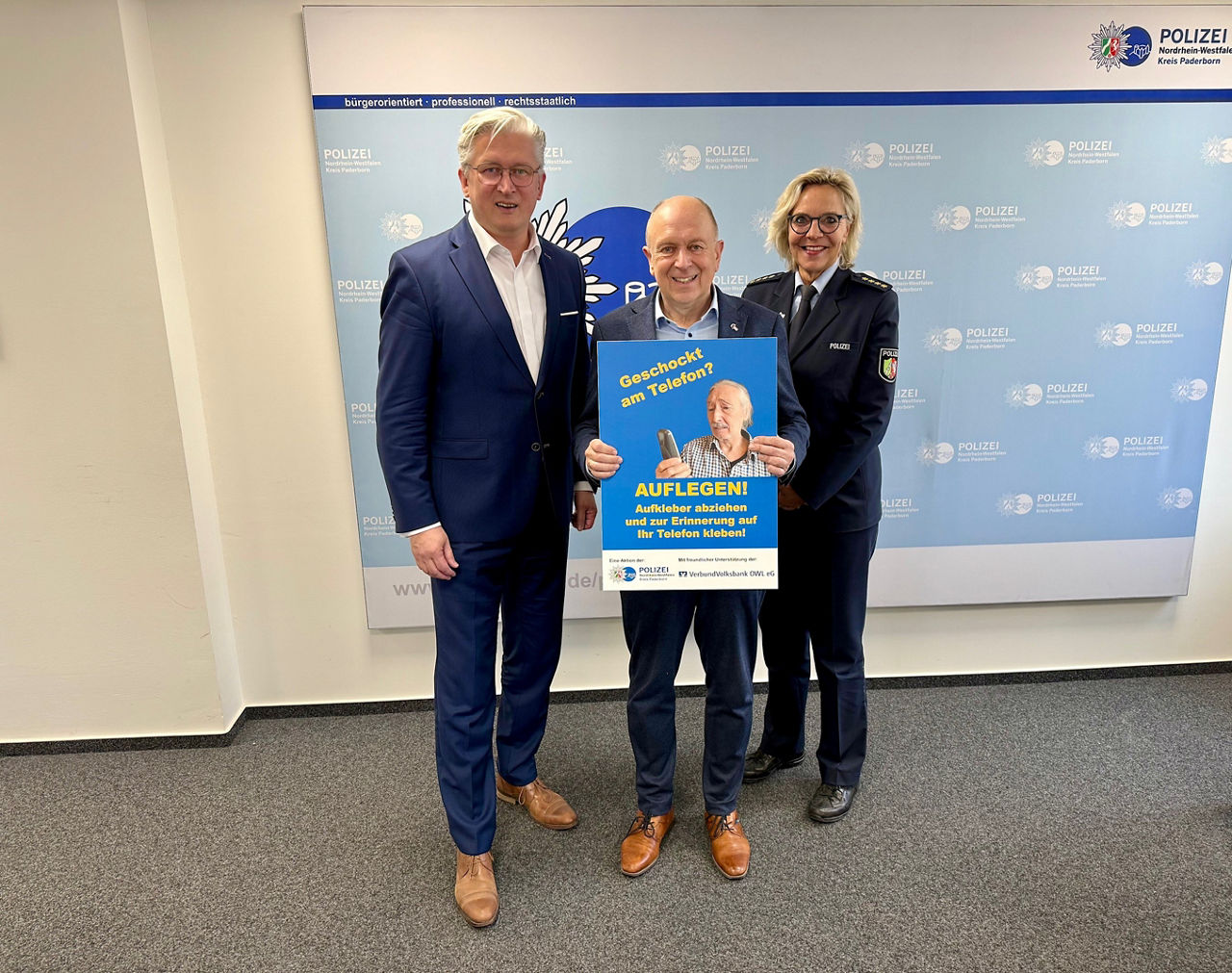 Bankvorstand Ansgar Käter und zwei Vertreter der Polizei mit einem Schild zur Aktion „Geschockt am Telefon? – Auflegen!“ 