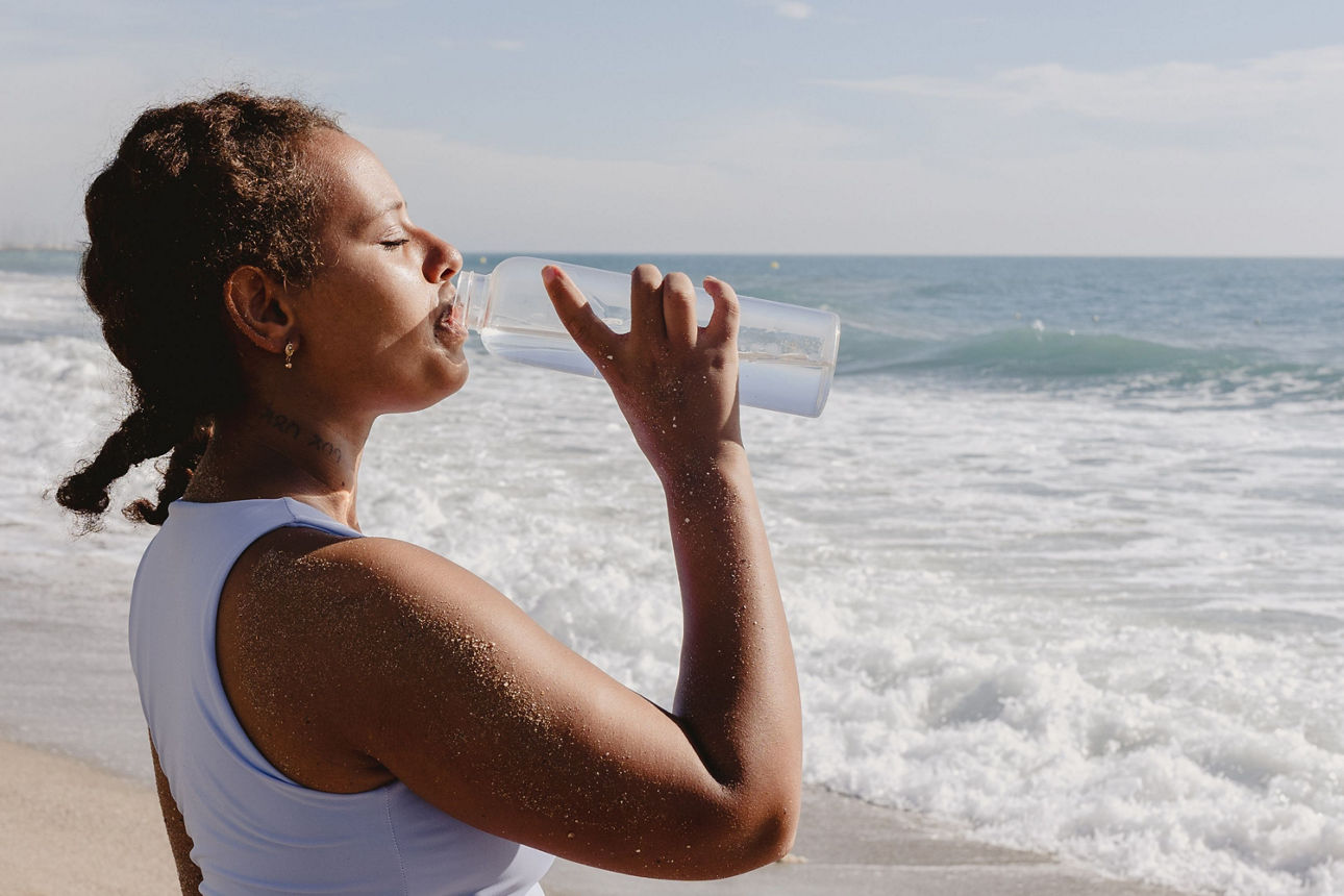 Eine Frau steht am Strand und trinkt aus einer Wasserflasche