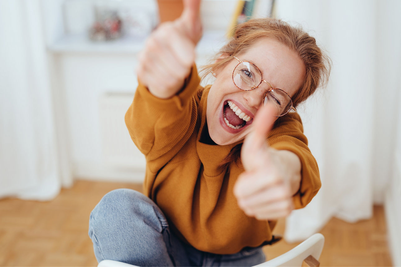 Happy pretty girl in glasses showing thumbs up gestures, sitting on chair in the room. Close-up portrait from high angle