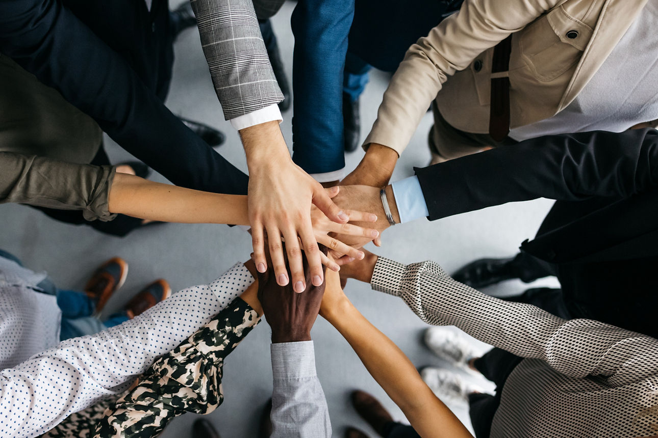 A high-angle shot of a group of male and female colleagues putting their hands together in an office. They are dressed in fashionable business clothes. Their faces are not visible, only their arms. Horizontal daylight indoor photo.