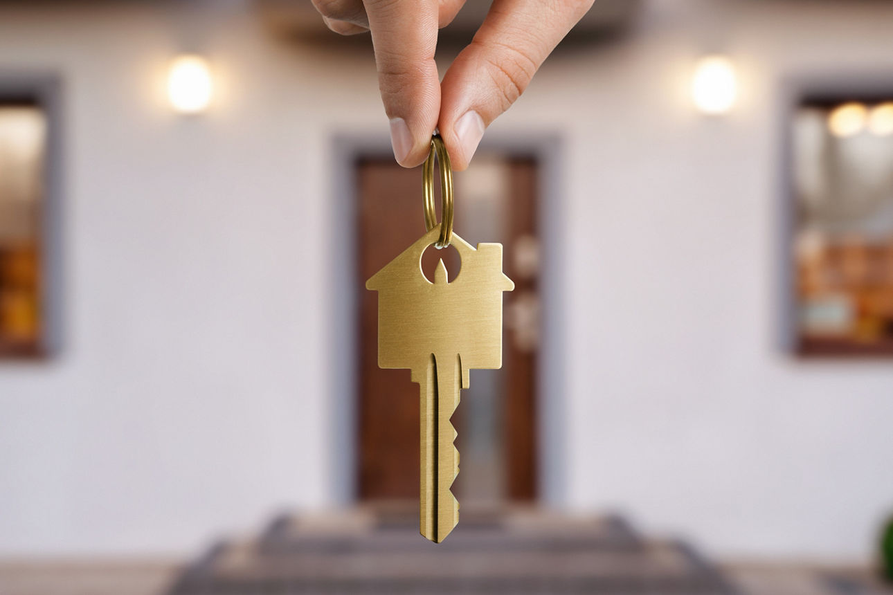 Eine Hand hält von oben einen Schlüssel ins Bild, der HIntergrund ist verschwommen und man erkennt ein Haus.