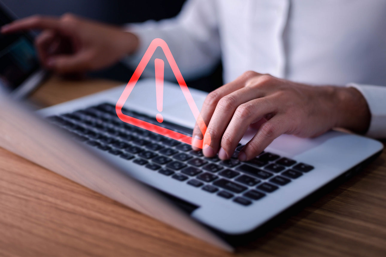 Hände auf einer Laptop-Tastatur. Darüber schwebt ein Warnzeichen.