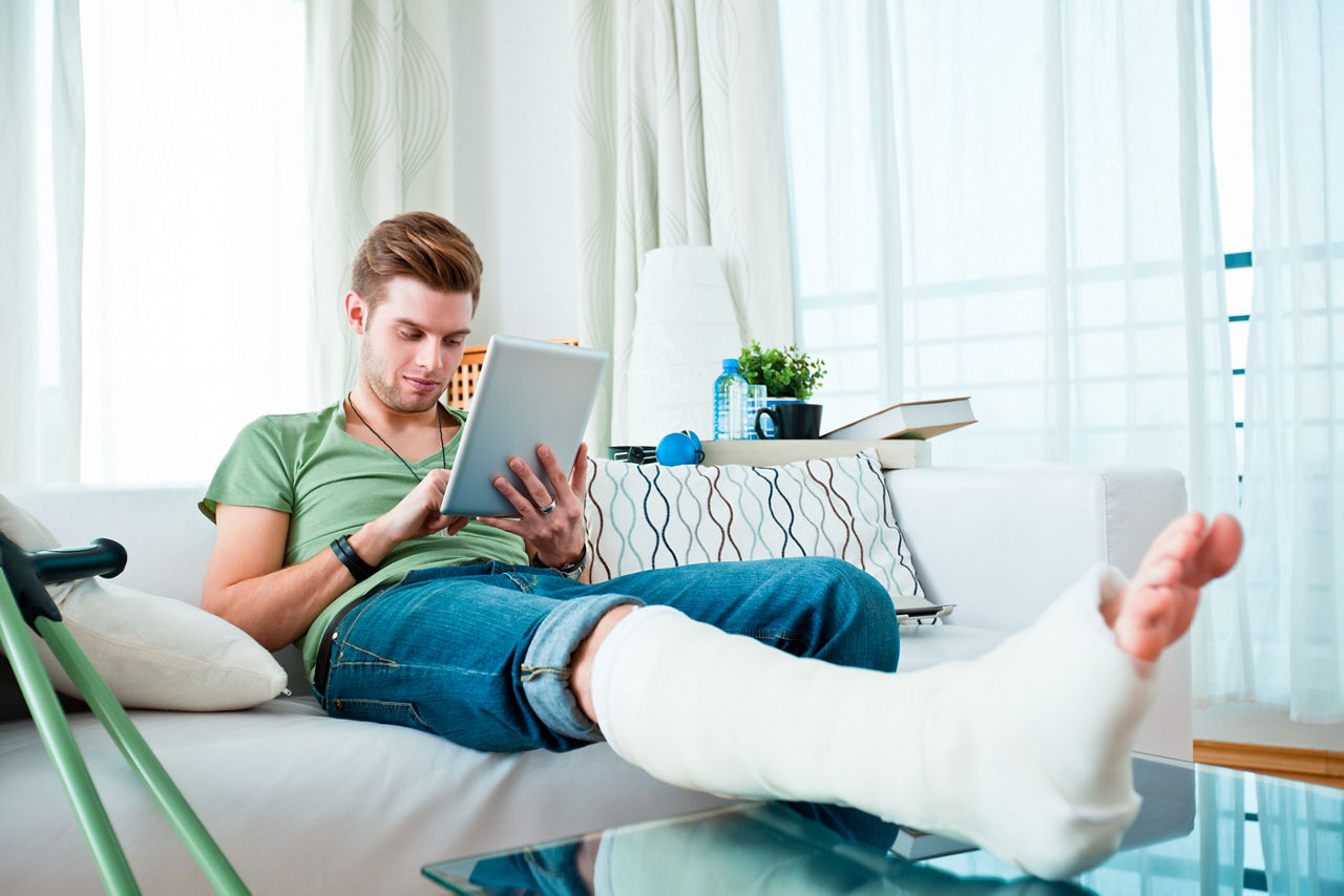 Young man with broken leg sitting on sofa at home and using digital tablet. 
