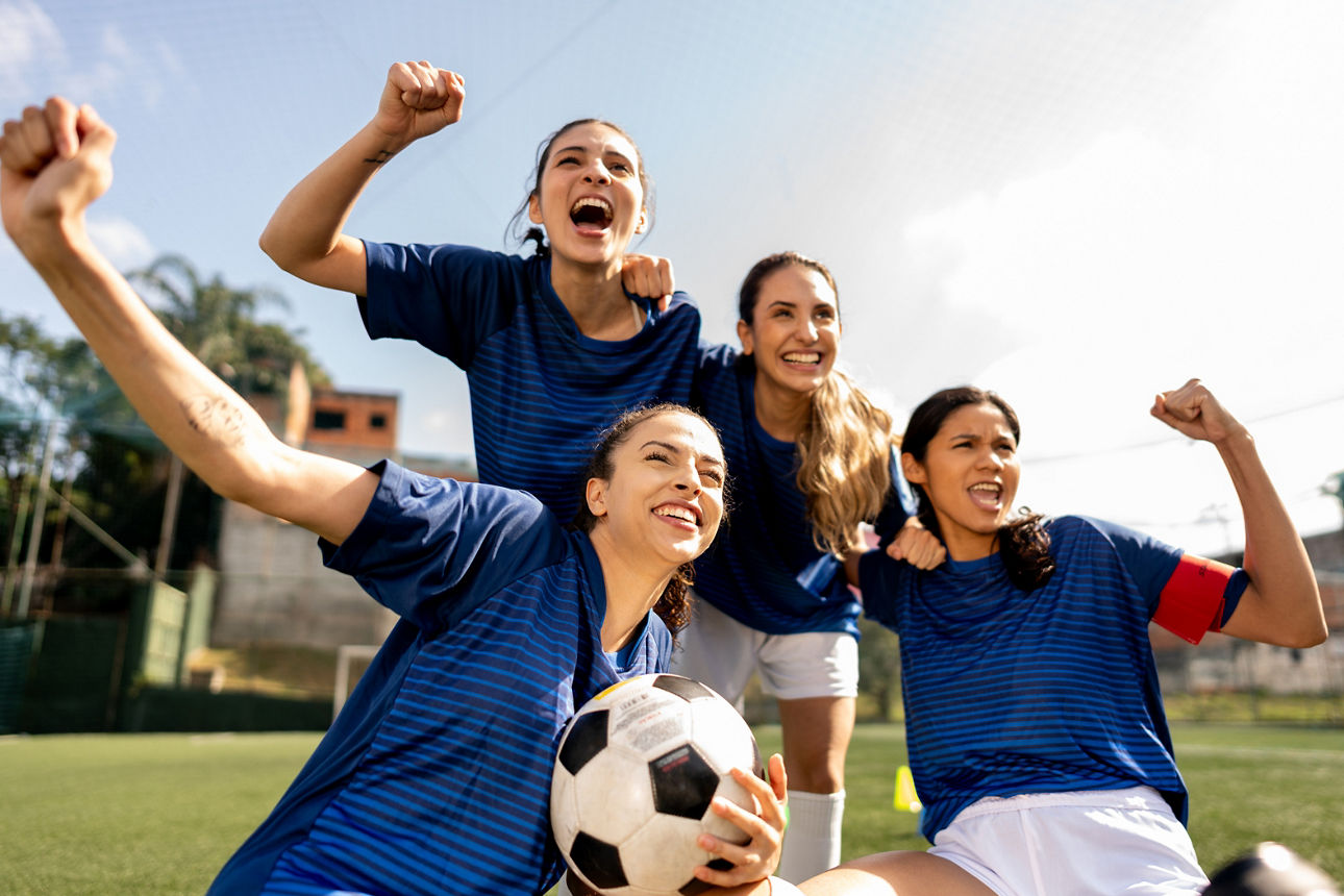 Vier junge Fußballspielerinnen mit einem Fußball jubeln auf einem Fußballfeld