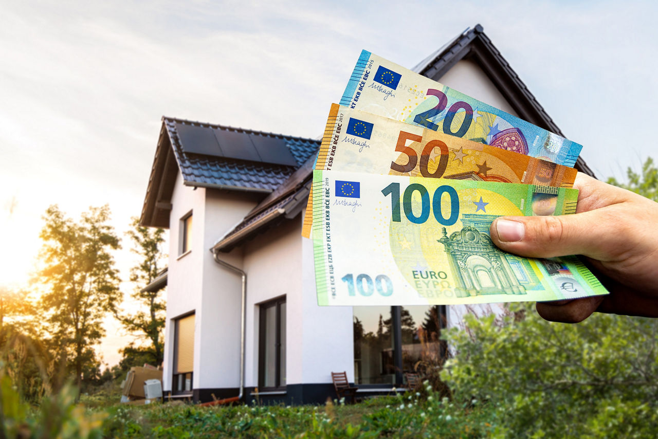 Eine Hand hält mehrere Euroscheine, im Hintergrund ist ein modernes Einfamilienhaus zu sehen.