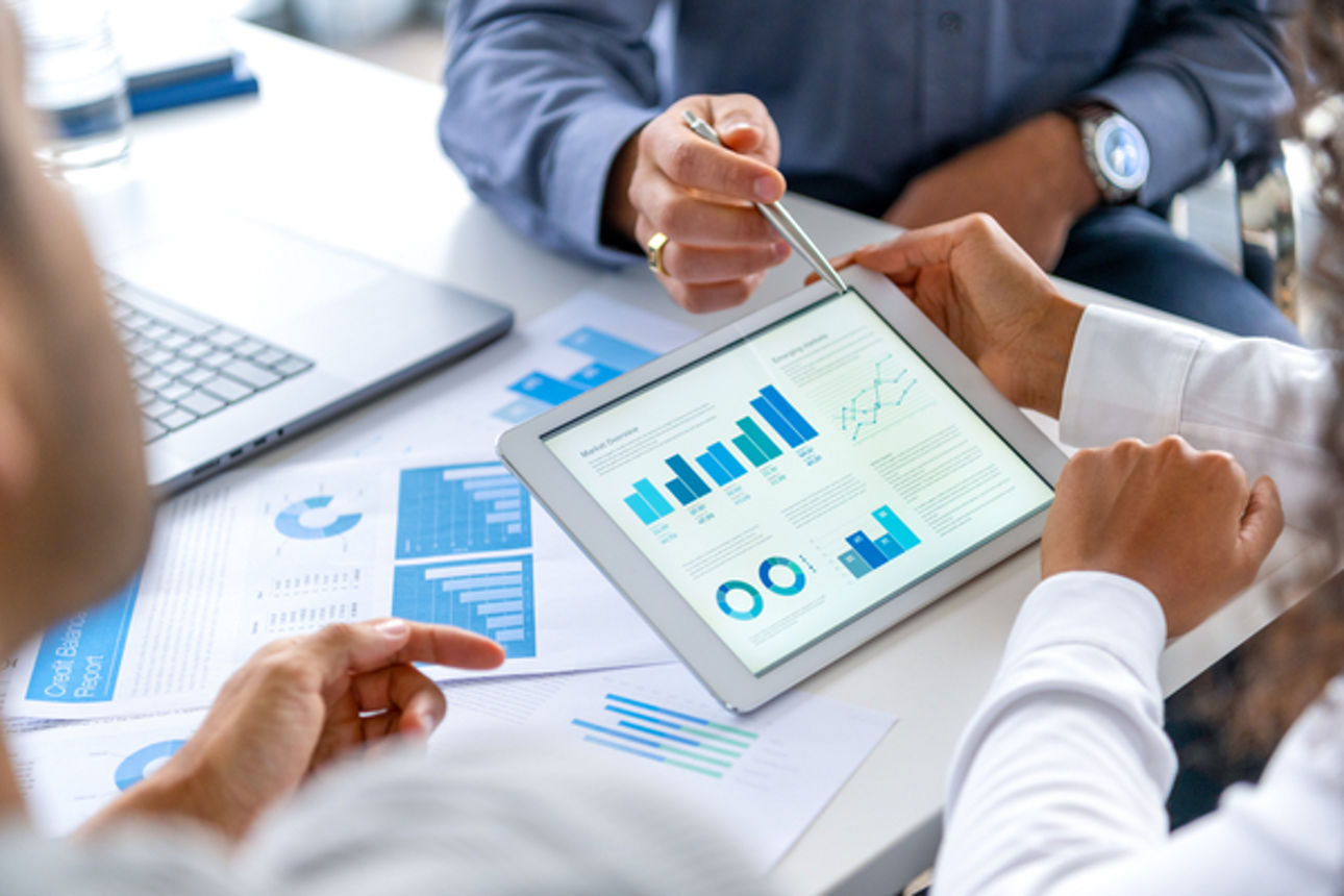 Close up of three people looking at financial data with graphs and charts.  All their hands can be seen and one person is pointing with a pen. There is paperwork on the desk showing more finance information