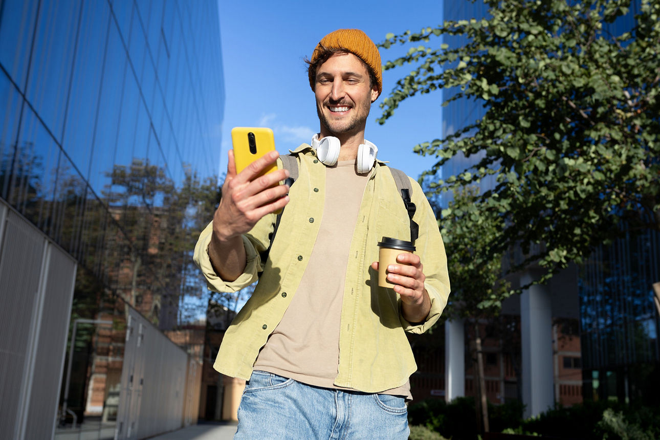 Young man with headphones and backpack using smartphone and drinking coffee while walking outdoors