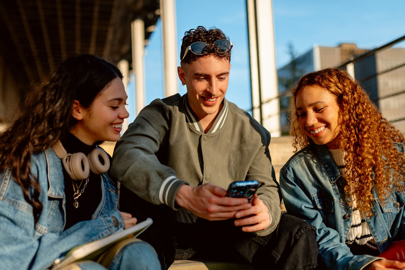 Three cheerful university students are sitting on steps, sharing a smartphone and enjoying their time together