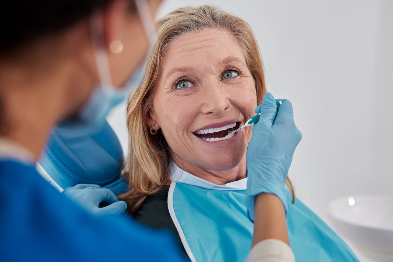 Close up of smiling old woman receiving dental cleaning with professional care. Female dentist using interdental tooth brush tool during oral hygiene session at clinic. Dental care provided during the medical visit, regular dentistry check up for maintaining oral health.