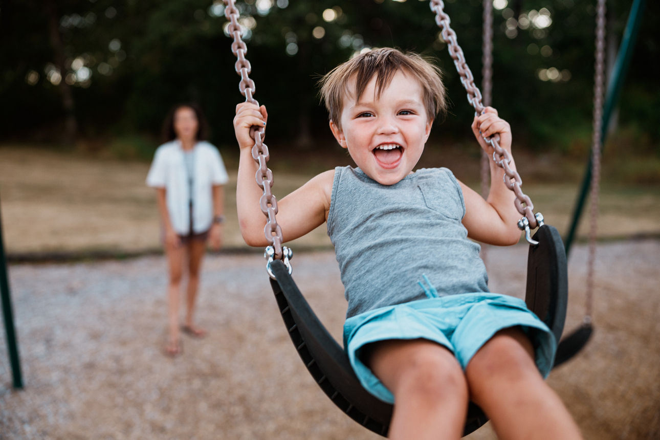 A three-year-old Eurasian boy laughs as his mom pushes him on a swing at the park.