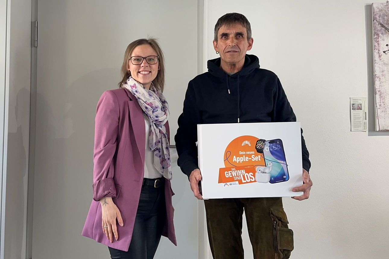 Es steht in einer Bankfiliale, eine Vermögensberaterin der VR-Bank und der Gewinner des Appel-Set. Der Gewinner hält das Set in den Händen, beide lächeln in die Kamera.  