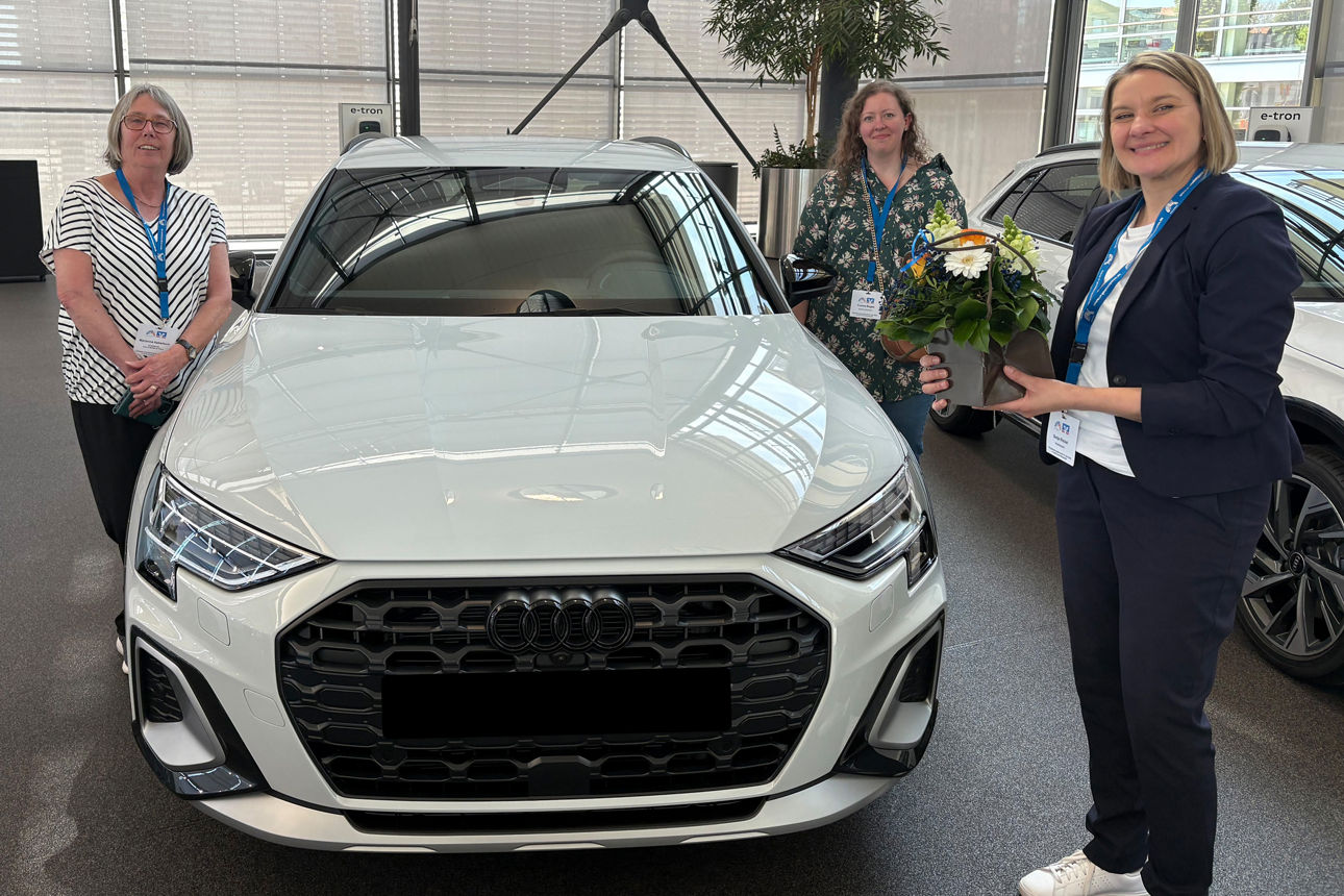 Drei Personen stehen im Autohaus vor einem weißen Audi; rechts hält jemand ein Blumengesteck.