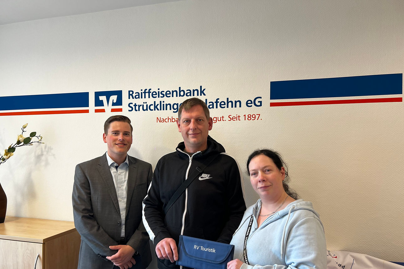 Kundenberater Tilman Winkler mit den Eheleuten Frobel stehend vor einer Wand mit dem Raibalogo.