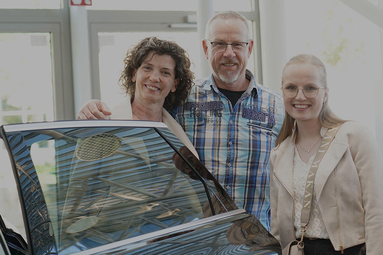 Zwei Frauen und ein Mann lächeln in die Kamera. Sie stehen in einer Autotür