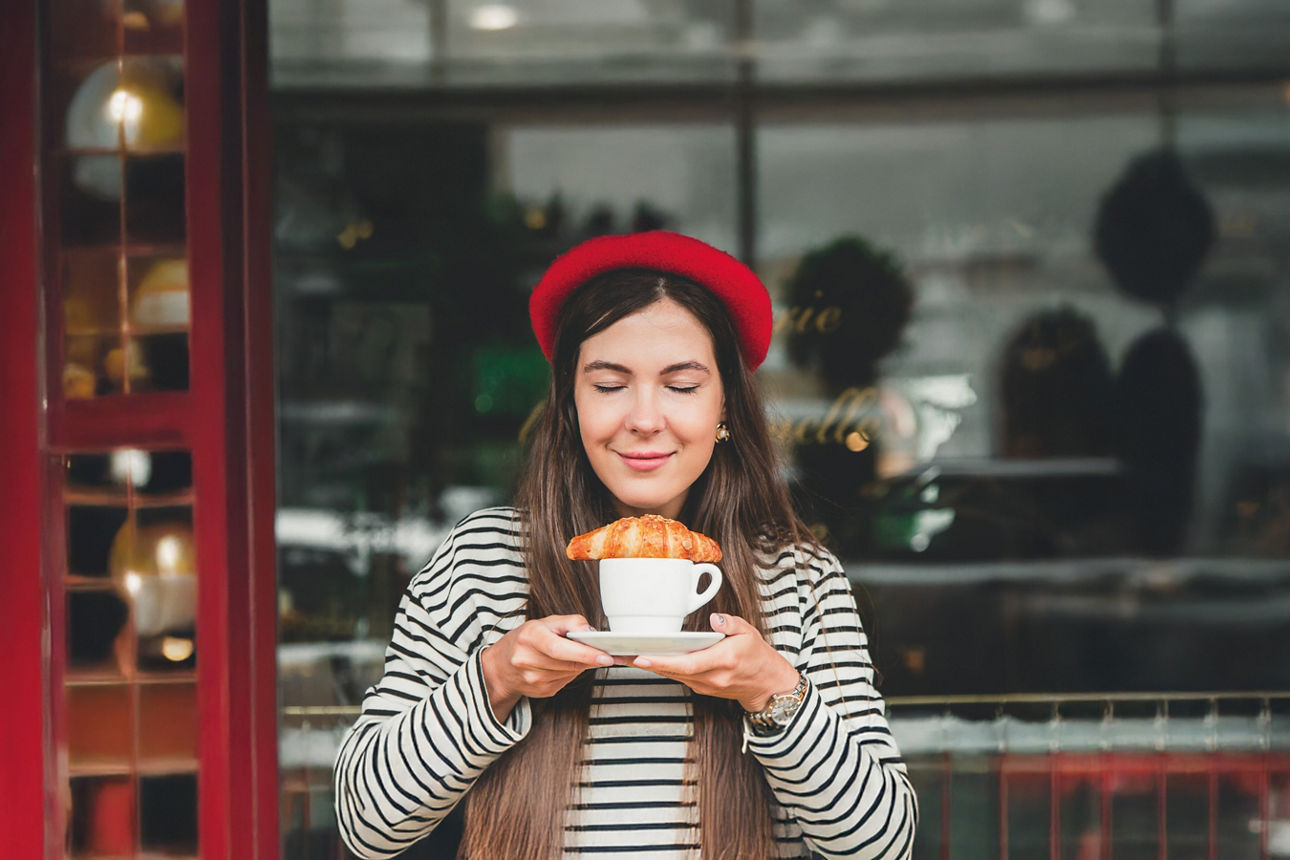 Eine Frau vor einem Café mit einer Tasse und einem Croissant in der Hand 