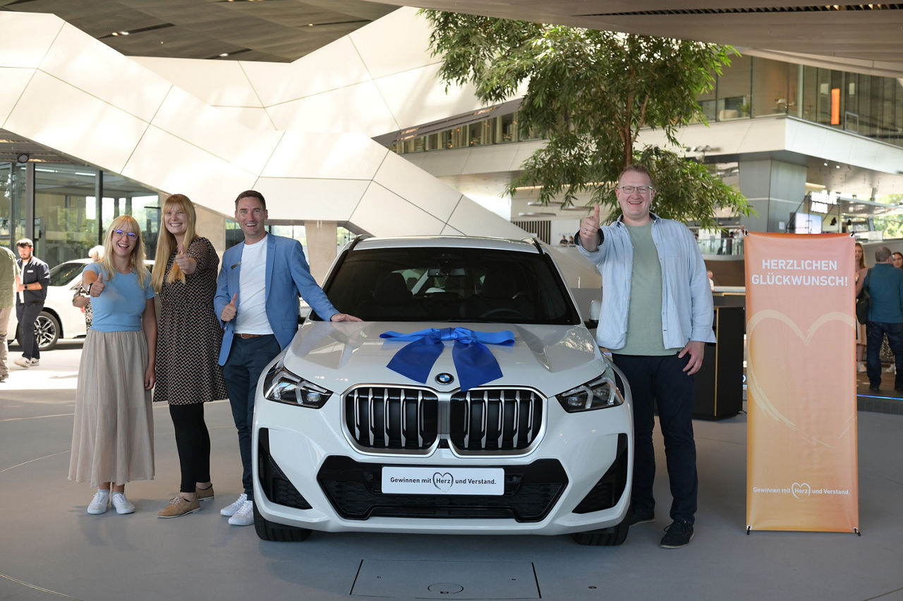 Das Bild zeigt die Vermögensberaterin und die Regionalleiterin der VR-Bank zusammen mit dem glücklichen Gewinner des BMW X1. Die Personen stehen links und rechts neben dem Neuwagen. Das Foto wurde in der BMW-Welt in München aufgenommen.