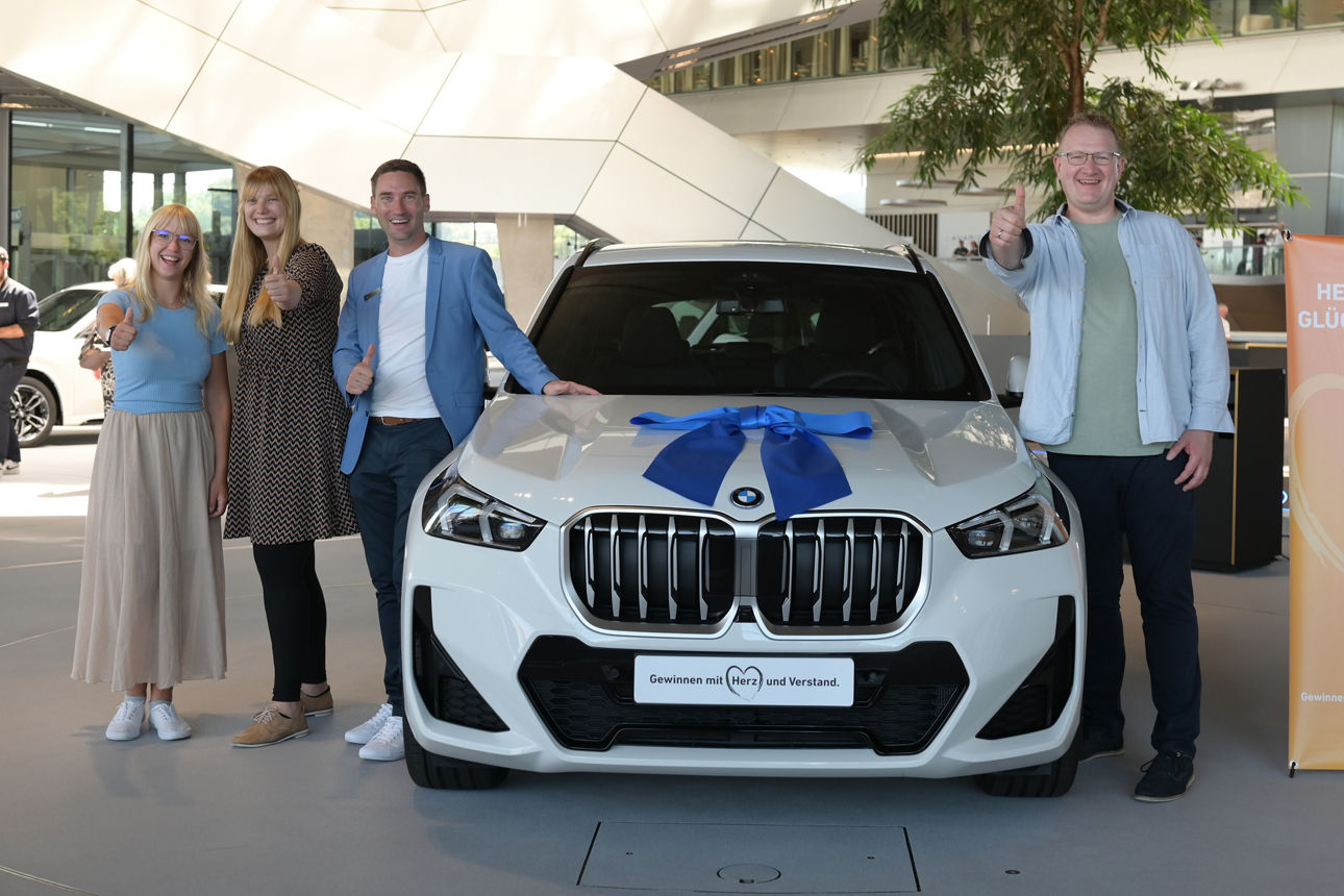 Das Bild zeigt die Vermögensberaterin und die Regionalleiterin der VR-Bank zusammen mit dem glücklichen Gewinner des BMW X1. Die Personen stehen links und rechts neben dem Neuwagen. Das Foto wurde in der BMW-Welt in München aufgenommen.