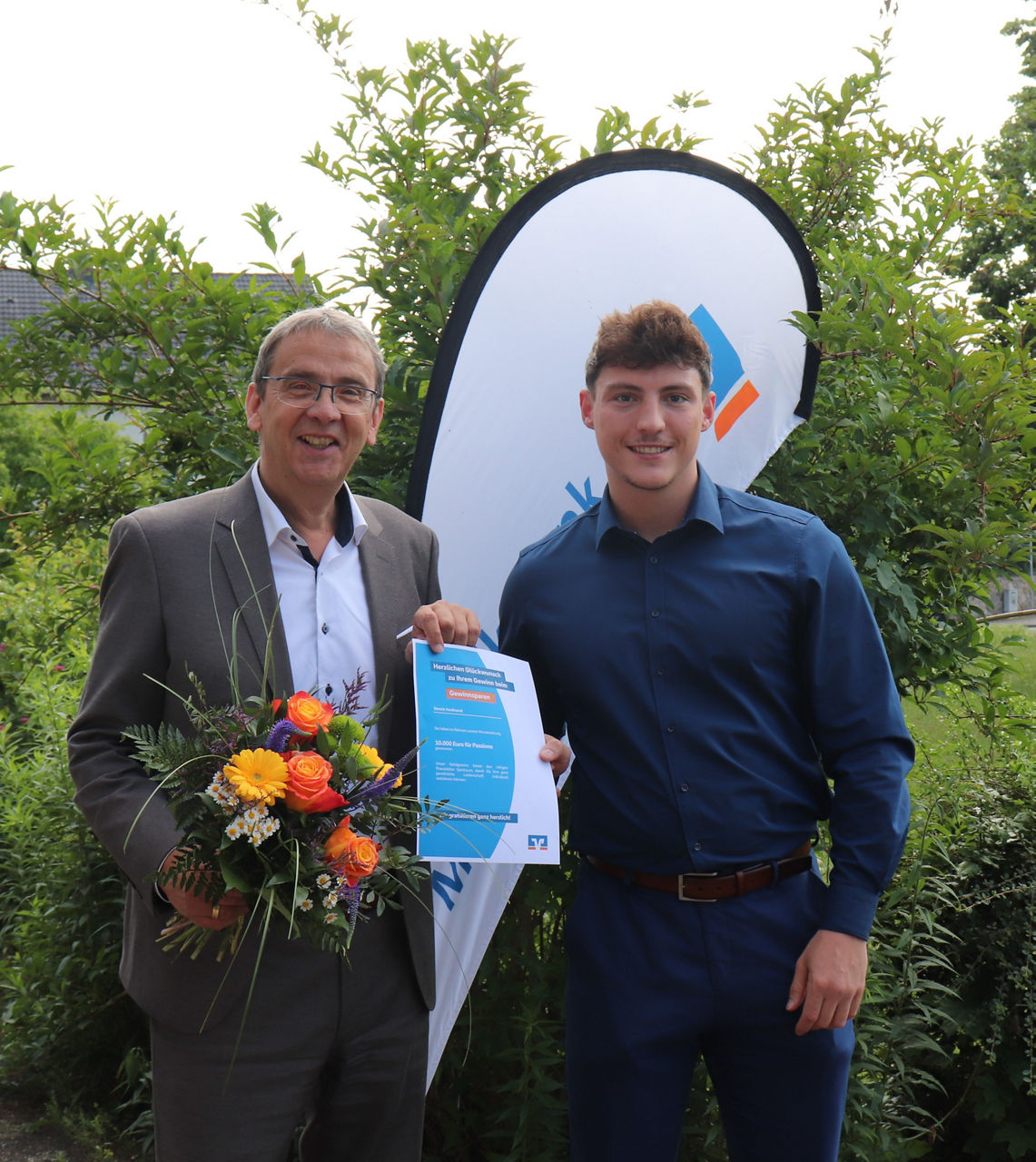 Voba Vorstand Herr Haas überreicht dem Gewinnspargewinner Herr Ferdinand symbolisch seinen Gewinn und einen Blumenstrauß