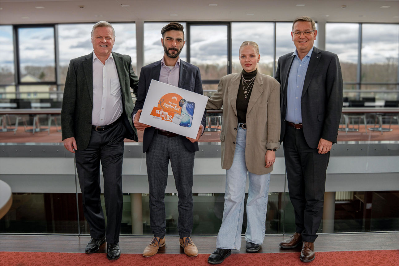 Eine junge Frau und drei Herren in Businesskleidung stehen nebeneinander in modernen Räumlichkeiten. Ein Herr hält einen Gewinn-Karton eines Apple-Sets in Händen 