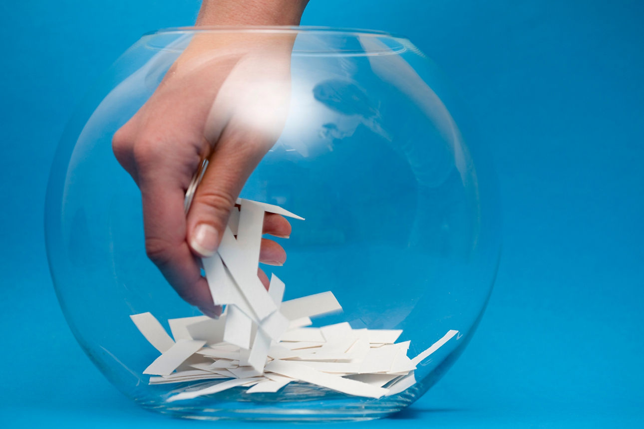 Hand zieht einen Loszettel aus einer Glasschale vor blauem Hintergrund – Symbol für Gewinnspiel oder Verlosung.