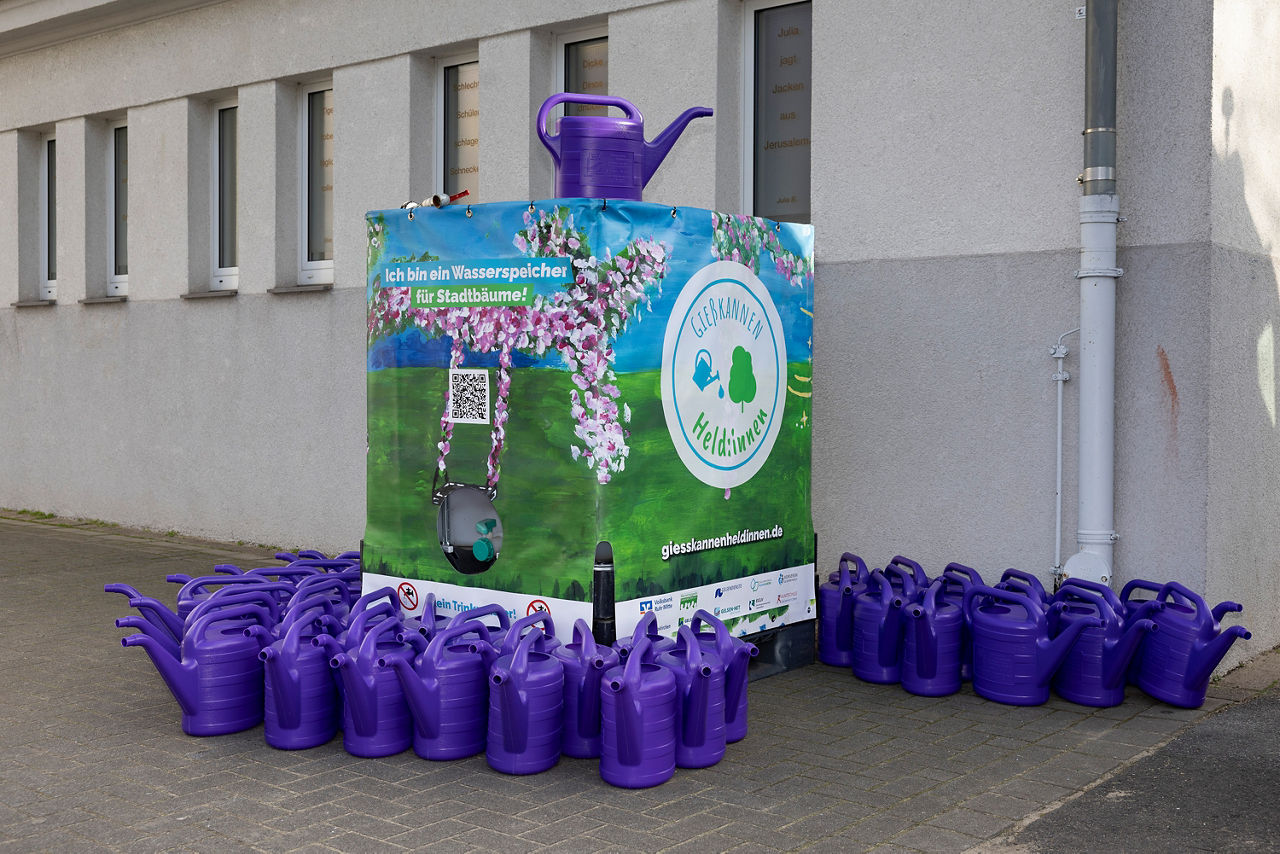 Wassertank für Regenwasser am Rande eines Schulhofes, am Boden umrandet von zahlreichen lila Gießkannen.