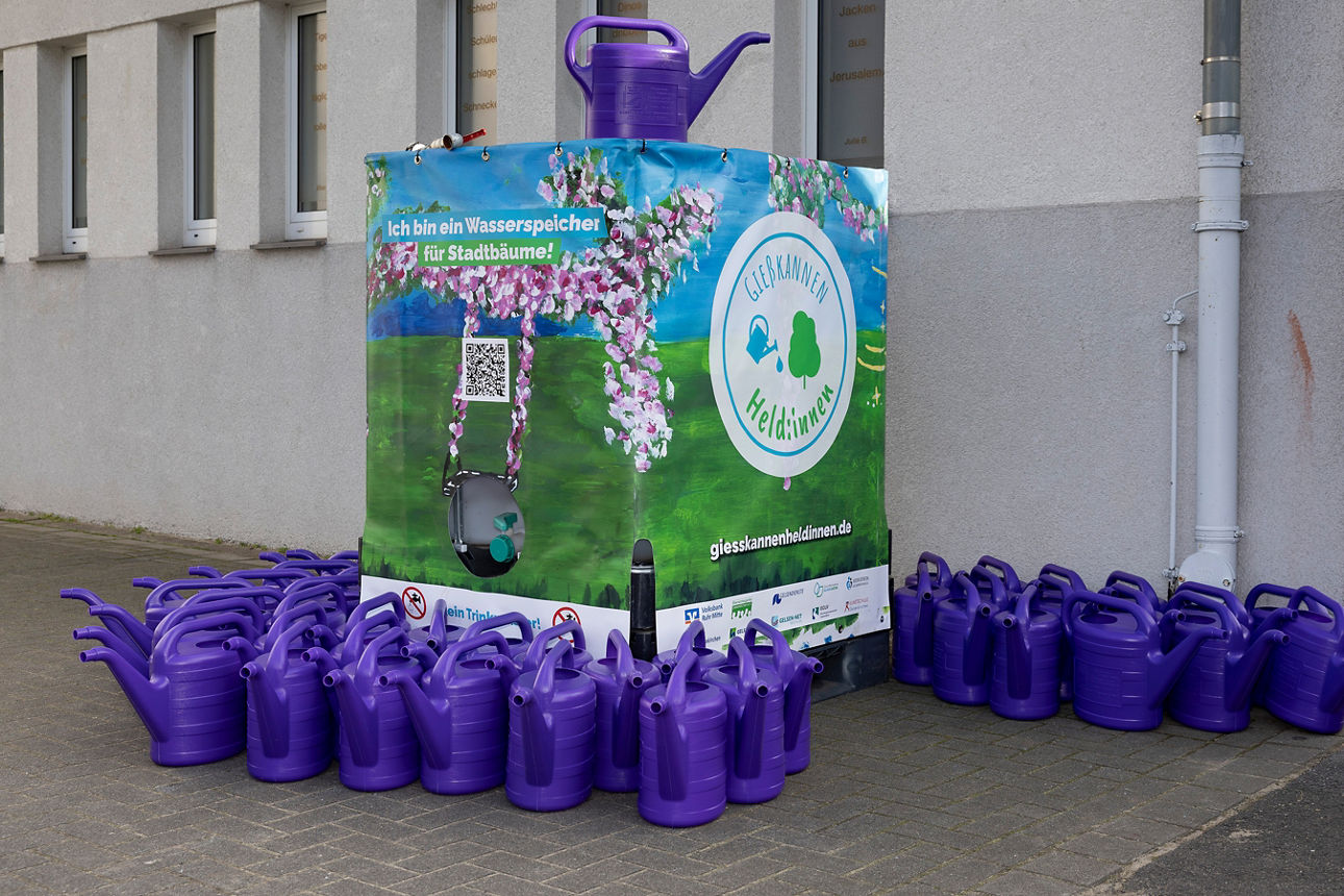 Wassertank für Regenwasser am Rande eines Schulhofes, am Boden umrandet von zahlreichen lila Gießkannen.