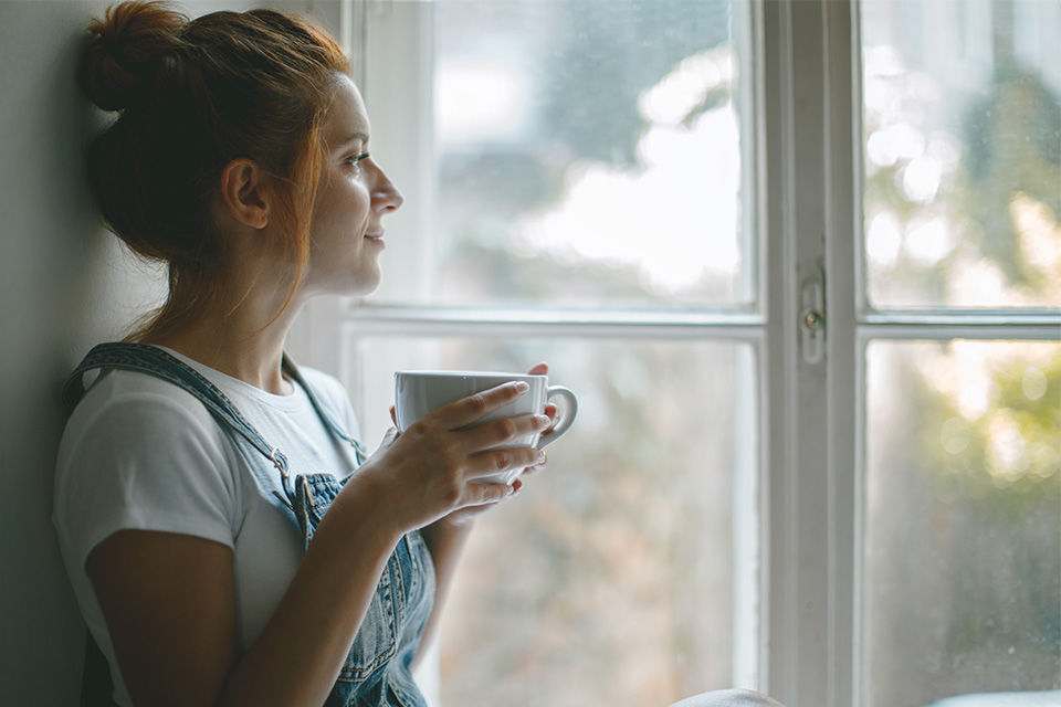 Eine Frau hält eine Tasse in den Händen und schaut aus dem Fenster