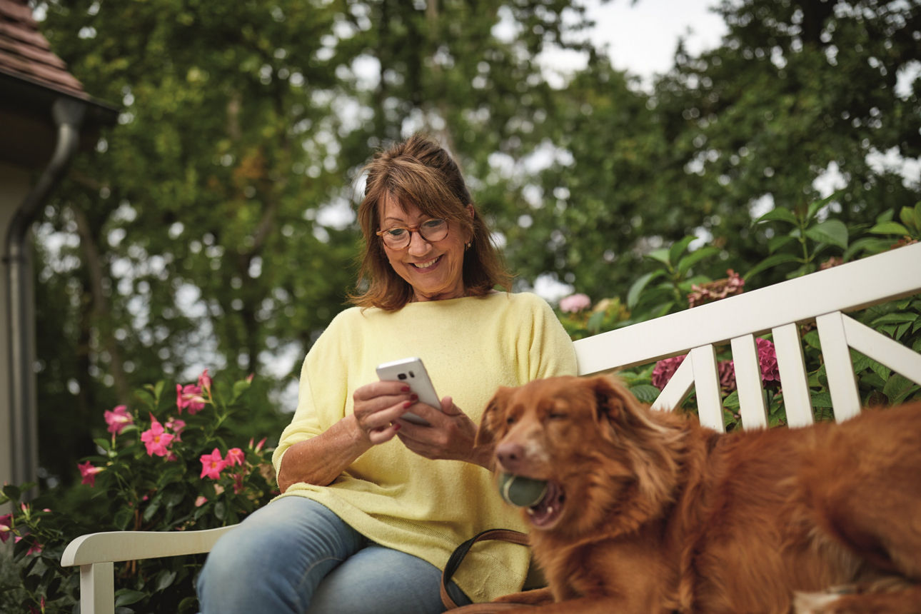Eine Frau sitzt mit ihrem Hund auf einer Gartenbank und checkt ihre Finanzen auf dem Handy.