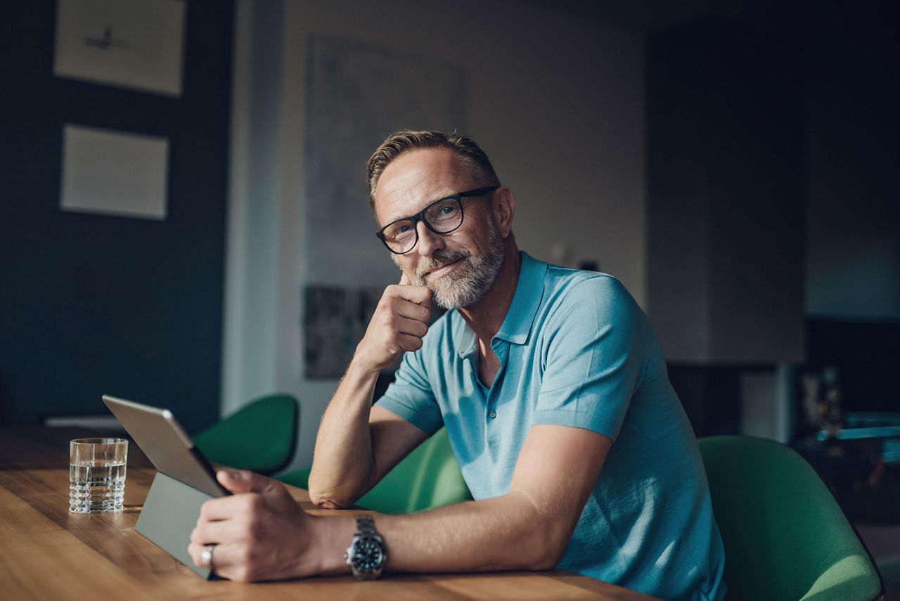 Mann mit Brille sitzt mit Tablet am Schreibtisch und guckt in die Kamera