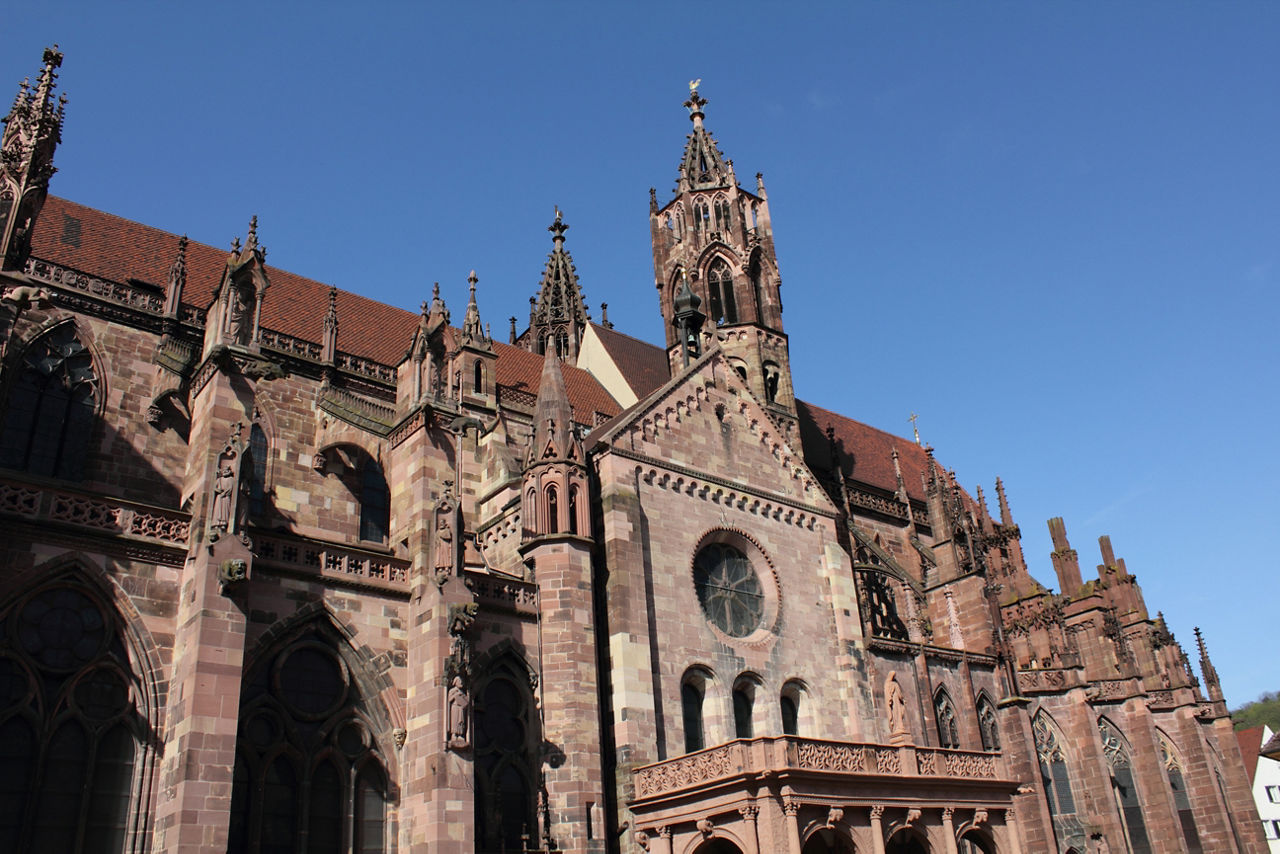 Das Bild zeigt eine imposante Kirche mit reich verzierten Türmen, Spitzbogenfenstern und zahlreichen Figurenfassaden im gotischen Stil vor blauem Himmel. Die Fassade besteht aus rötlich-braunem Naturstein mit einem großen Rundfenster in der Mitte.