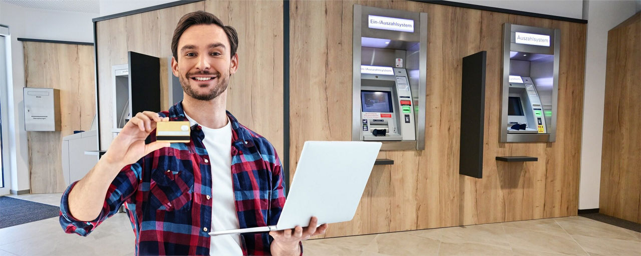 Kunde steht im SB-Raum einer Bank mit einer Kreditkarte und einem Laptop in der Hand 