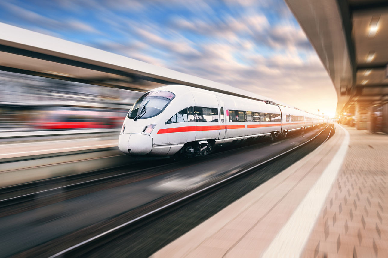 Weißer moderner Hochgeschwindigkeitszug während der Fahrt durch einen Bahnhof bei Sonnenuntergang