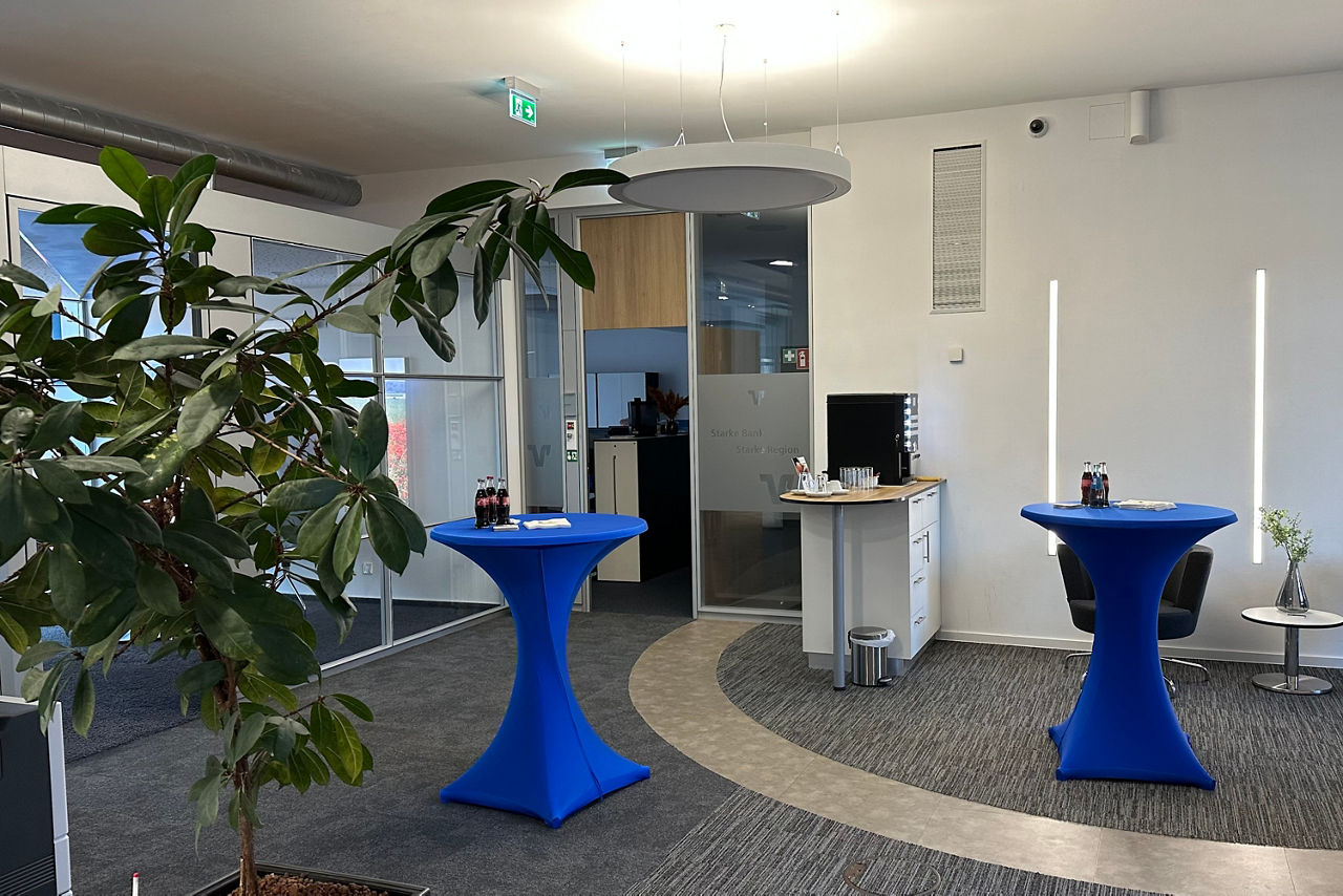 Büro mit zwei hohen Stehtischen mit blauen Hussen, großem grünen Pflanzenzweig links und kleinem Empfangstresen im Hintergrund