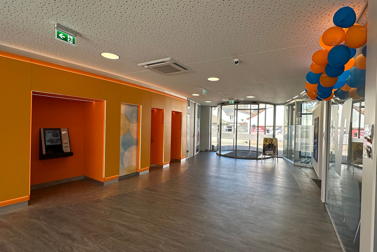 Flur mit orangen Wänden, mehreren Nischen, Glasdrehtür am Ende und blauen sowie orangen Luftballons an der Decke rechts