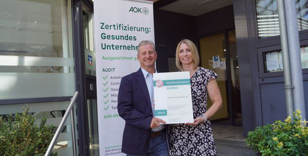 Ein Mann und eine Frau stehen vor einem Gebäude und halten ein Zertifikat mit der Aufschrift „Zertifizierung: Gesundes Untern