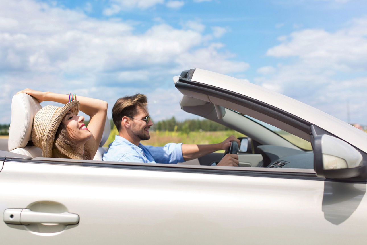Mann fährt in einem offenen Cabrio durch eine sommerliche Landschaft. Neben ihm sitzt entspannt eine Frau mit Sommerhut. 