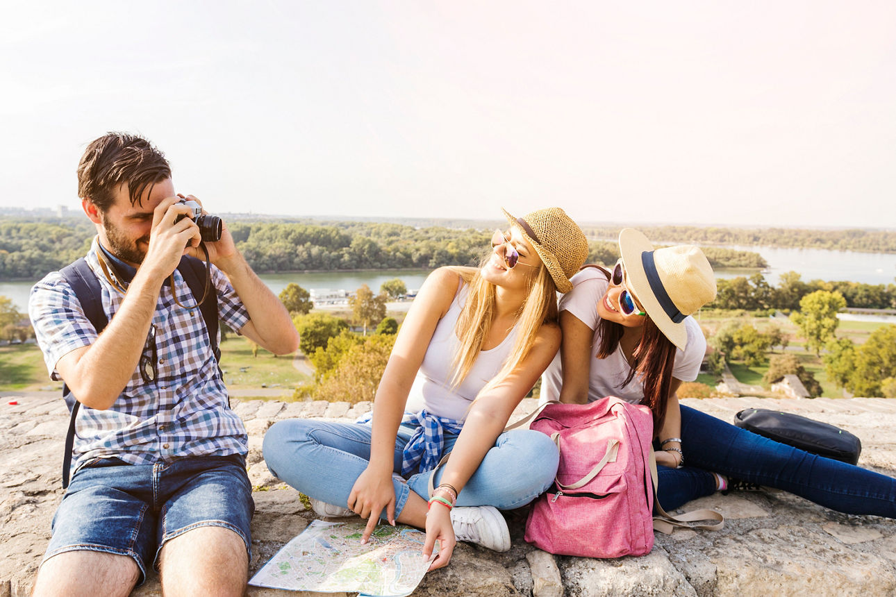 Drei junge Reisende sitzen auf einem Felsen mit Blick ins Tal, zwei lachen mit Sonnenhüten, einer fotografiert die Aussicht.