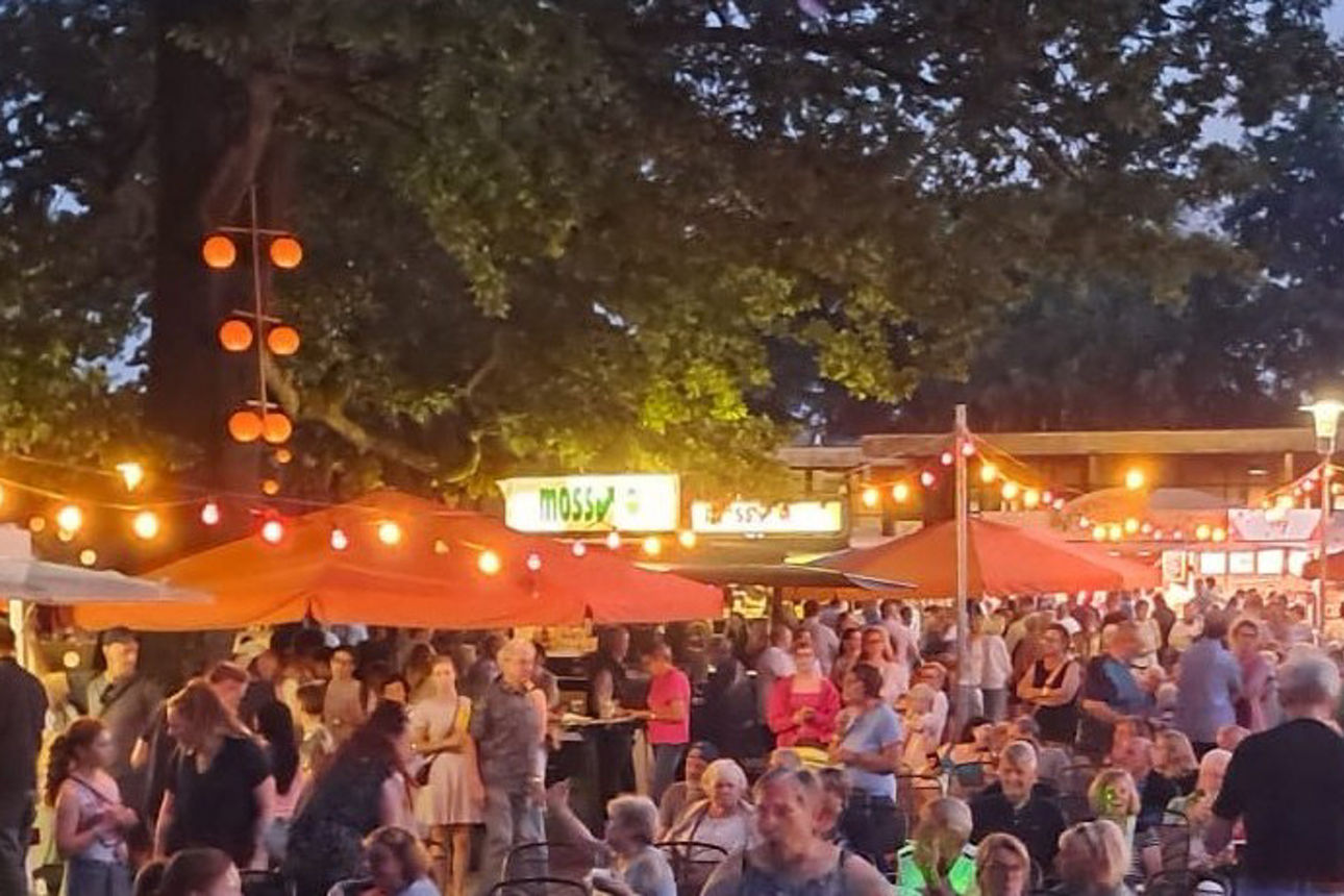 Zahlreiche Menschen besuchen eine Veranstaltung im Freien mit beleuchteten Marktständen und Sitzbereichen unter großen Bäumen