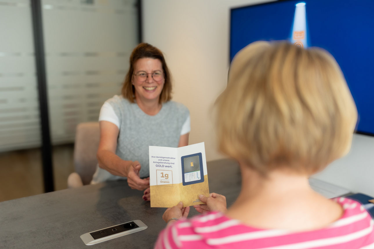 Eine Beraterin sitzt mit einer Kundin am Beratungstisch und überreicht ihr einen Gramm Gold. 