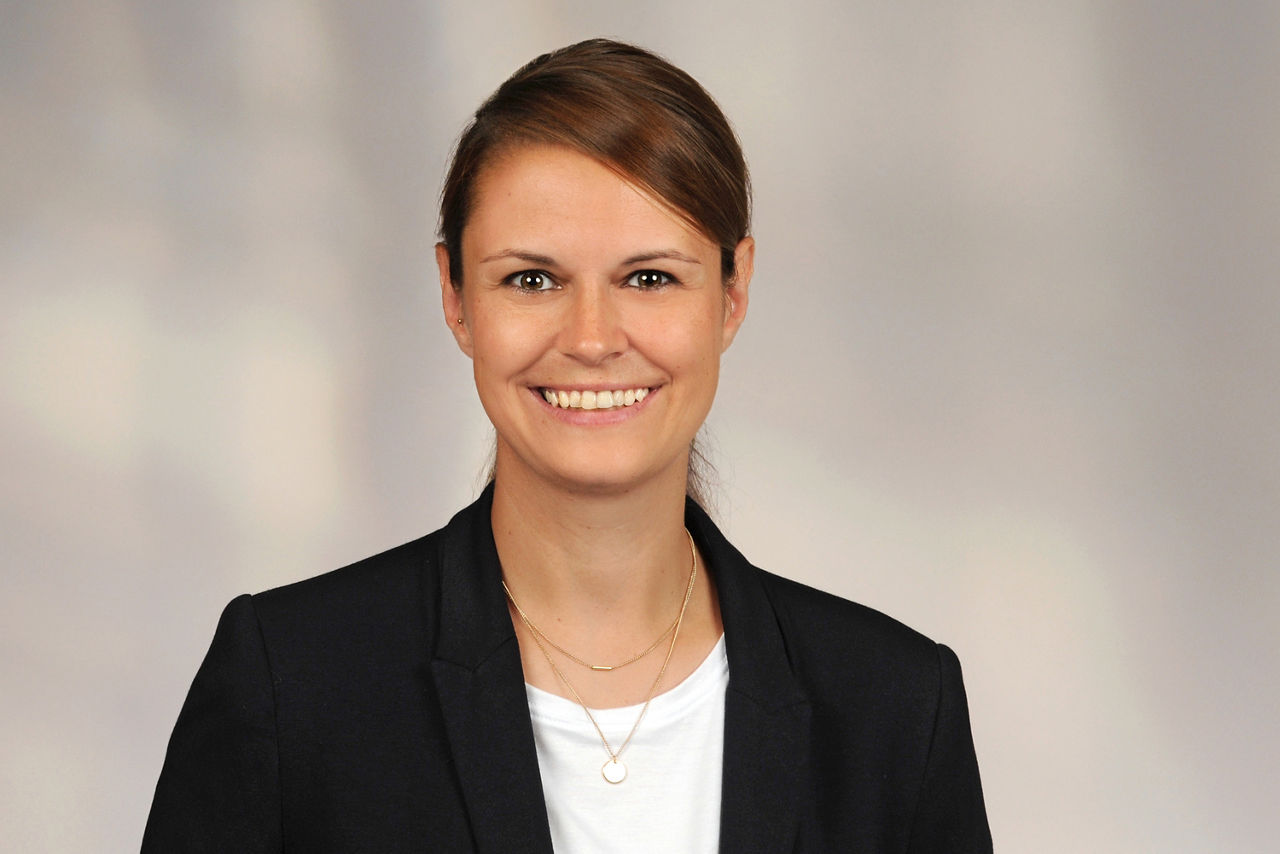 Yvonne Grümmer hat braune Haare und einen Seitenscheitel. Die Haare sind zu einem Zopf gebunden. Sie trägt ein Businessoutfit