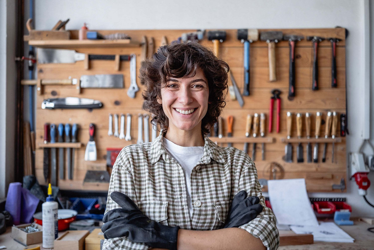 Portrait shot of a young Caucasian female craftsperson in plaid shirt, positioned against organized wall-mounted tool display in professional woodworking workshop.