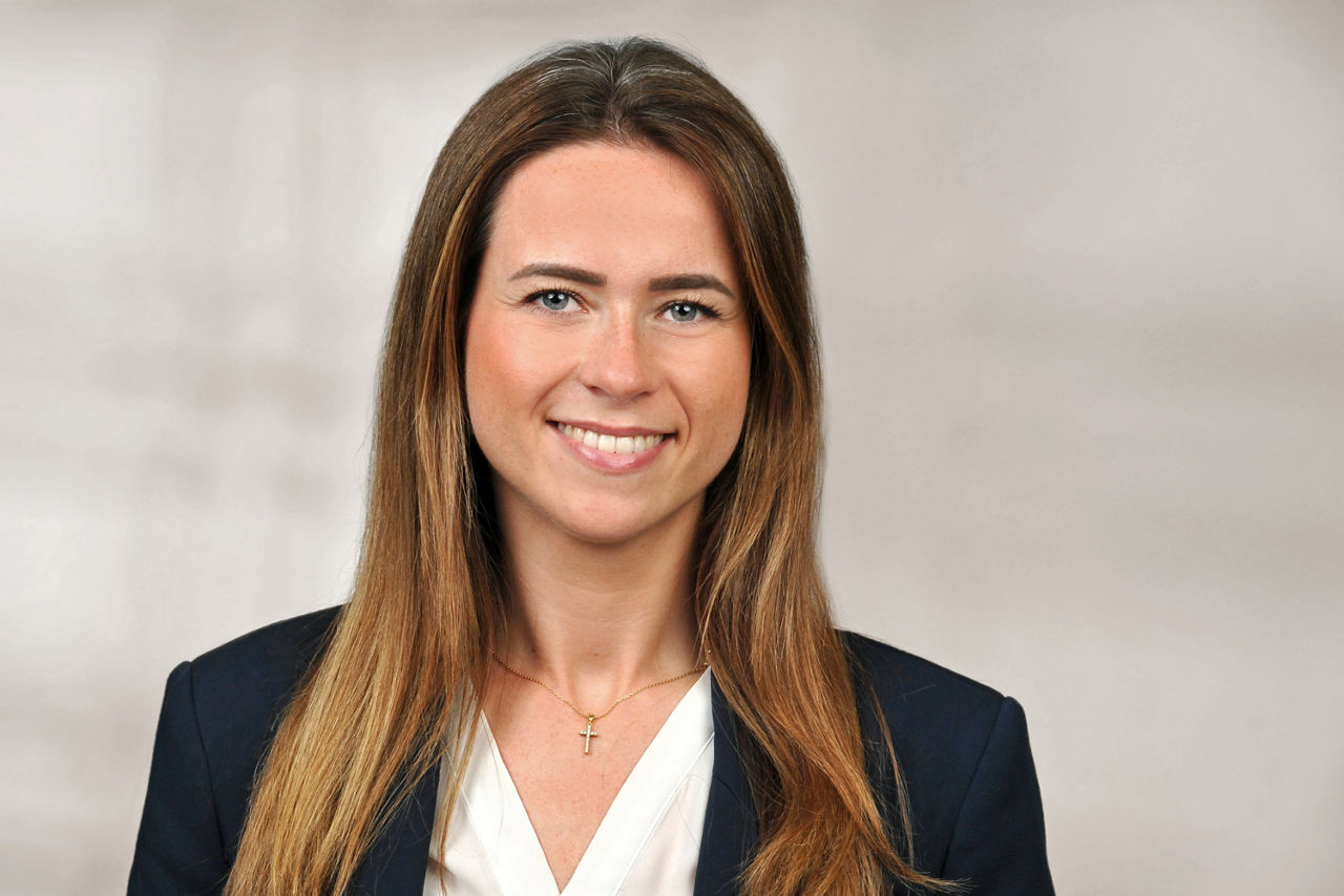 Frau mit langen braunen Haaren, trägt weißen Blusenkragen, schwarzen Blazer und eine Halskette mit Kreuzanhänger