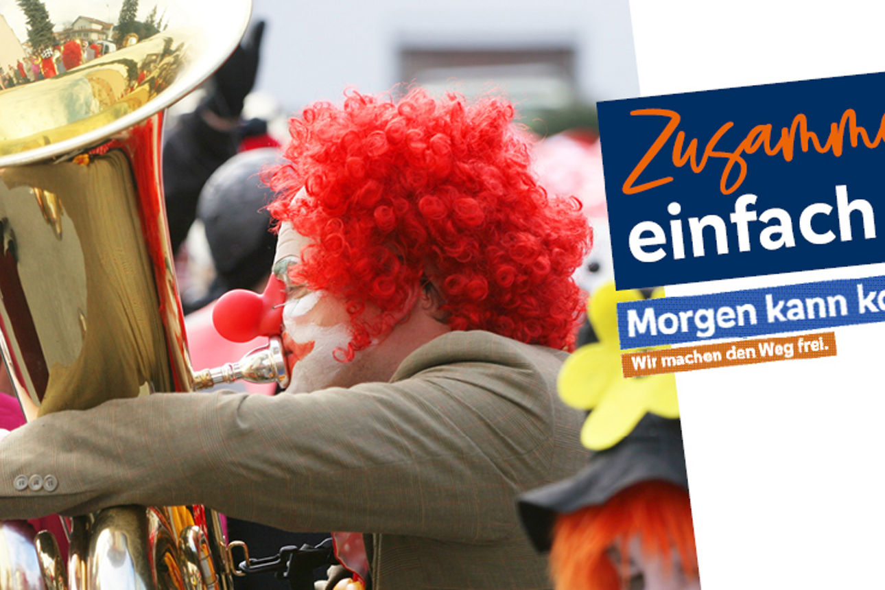 Person mit roter Clownperücke spielt Tuba und hält Notenblatt in der Hand